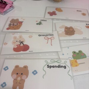 May include: A collection of clear plastic budget envelopes with cute cartoon illustrations. Each envelope is labeled with categories like "Groceries" and "Spending". The envelopes feature images of a bear, bunny, and frog.