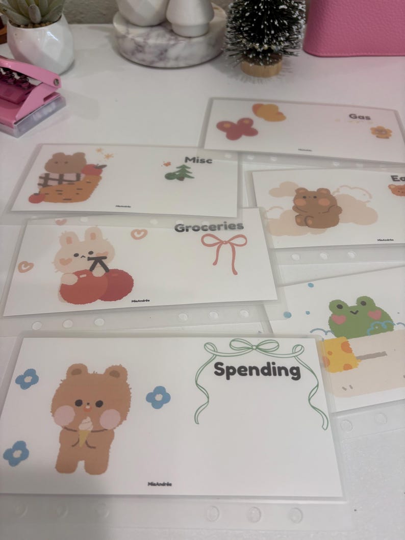 May include: A collection of clear plastic budget envelopes with cute cartoon illustrations. Each envelope is labeled with categories like "Groceries" and "Spending". The envelopes feature images of a bear, bunny, and frog.