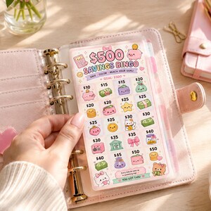 Puede incluir: Un juego de bingo de ahorro rosa con ilustraciones bonitas y el texto "$500 Savings Bingo". La tarjeta se sostiene en una carpeta rosa con detalles dorados. Un cuaderno y un bol&iacute;grafo rosas son visibles en el fondo.