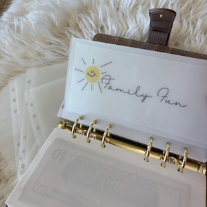 May include: A brown leather planner with gold rings, open to reveal a clear pocket with the words "Family Fun" and a smiling sun graphic. The planner contains several clear, lined pockets, and a glimpse of a ten-dollar bill.