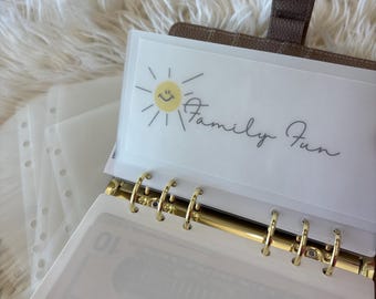 May include: A brown leather planner with gold rings, open to show a clear pocket with the words "Family Fun" and a smiling sun graphic. The planner contains clear, lined pockets, and a glimpse of a ten-dollar note.