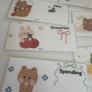 May include: White budget envelopes with cute illustrations of a bear, bunny, and other designs. Each envelope is labeled with categories like "Groceries" and "Spending". The envelopes are decorated with pastel colors and whimsical graphics.