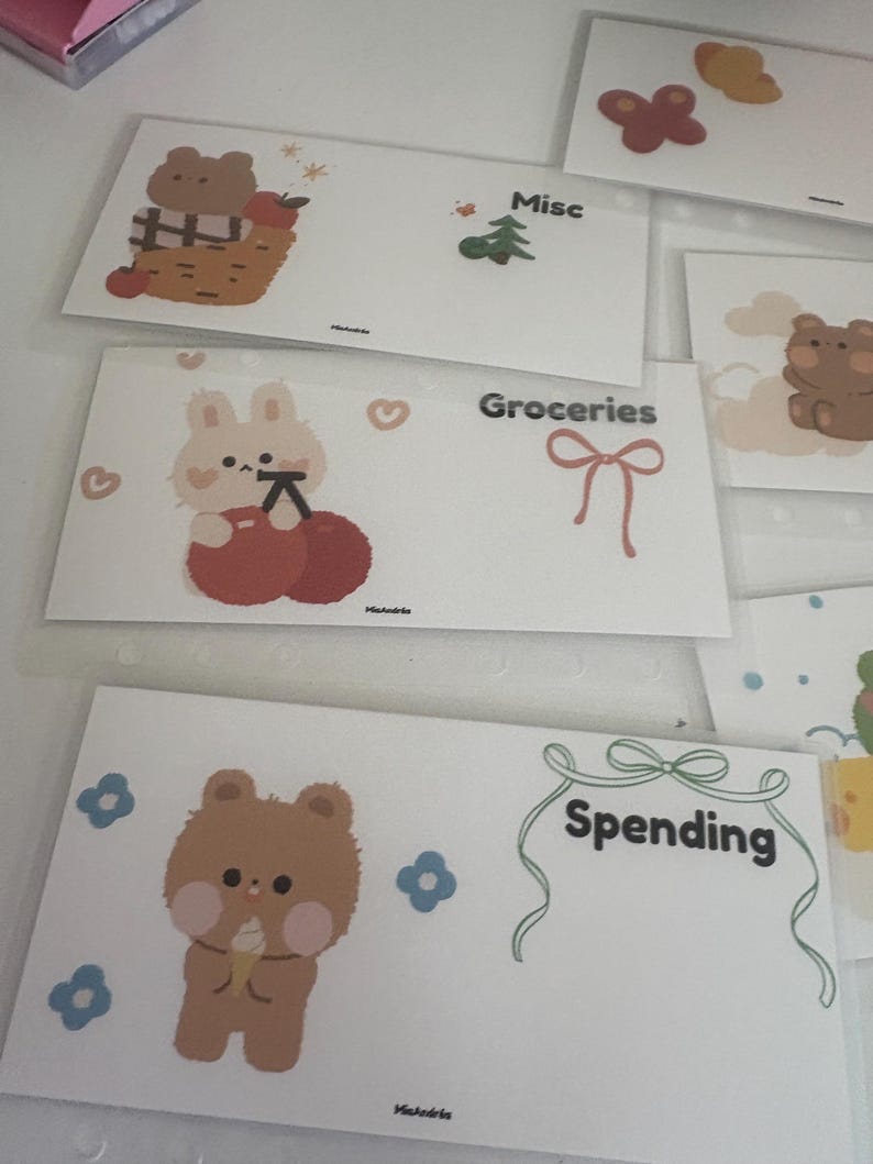 May include: White budget envelopes with cute illustrations of a bear, bunny, and other designs. Each envelope is labeled with categories like "Groceries" and "Spending". The envelopes are decorated with pastel colors and whimsical graphics.