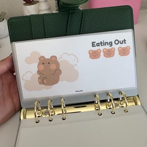 May include: A green leather binder with gold-colored rings, holding a clear plastic sleeve. The sleeve features the text "Eating Out" and illustrations of a bear and clouds. A pink binder is visible in the background.