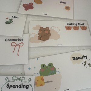 May include: A collection of white budget envelopes with various illustrations and labels. Designs include a butterfly, bear, frog, and bow. Labels include "Gas", "Eating Out", "Beauty", "Spending", "Groceries", and "Misc".