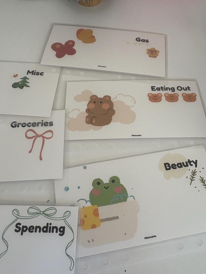 May include: A collection of white budget envelopes with various illustrations and labels. Designs include a butterfly, bear, frog, and bow. Labels include "Gas", "Eating Out", "Beauty", "Spending", "Groceries", and "Misc".