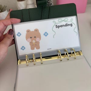May include: Open green leather binder with gold rings, holding a clear plastic sleeve. The sleeve features a cartoon bear illustration and the word "Spending" in green script. Blue flower graphics are also present.
