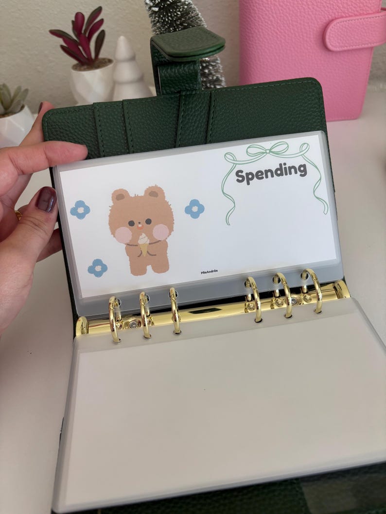 May include: Open green leather binder with gold rings, holding a clear plastic sleeve. The sleeve features a cartoon bear illustration and the word "Spending" in green script. Blue flower graphics are also present.