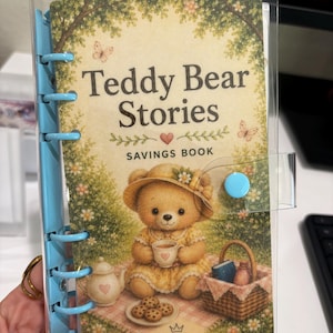 May include: A clear plastic binder with a light blue metal ring mechanism and a snap closure. The cover features a teddy bear in a straw hat having a picnic with tea and cookies, with the text Teddy Bear Stories SAVINGS BOOK and QUEEN DOLLAR.