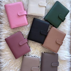 Op de afbeelding: Een verzameling leren persoonlijke planners in verschillende kleuren, waaronder roze, wit, groen, zwart, bruin en taupe. Elke planner heeft een veilige tabsluiting en ligt op een zachte witte ondergrond.