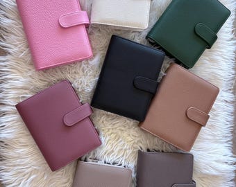 Puede incluir: Una colección de agendas personales con textura de cuero en varios colores, incluyendo rosa, blanco, verde, negro, marrón y topo. Cada agenda tiene un cierre de pestaña seguro y está dispuesta sobre una superficie blanca y esponjosa.