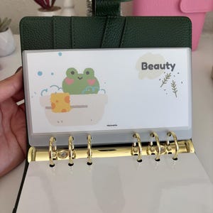 May include: Dark green binder with gold rings, open to reveal a white insert with a cartoon frog in a bathtub. The insert has the word "Beauty" and decorative elements. The binder is open, showing the interior.