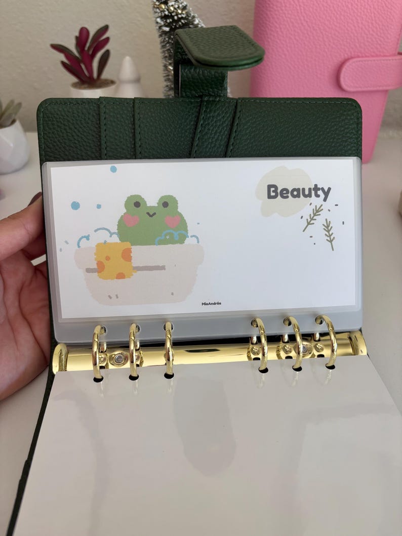 May include: Dark green binder with gold rings, open to reveal a white insert with a cartoon frog in a bathtub. The insert has the word "Beauty" and decorative elements. The binder is open, showing the interior.