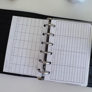 Op de afbeelding: Open zwarte planner met witte gelinieerde pagina's. De linkerpagina is een maandelijkse kalenderindeling en de rechterpagina is een wekelijkse planning. De planner is open om de pagina's weer te geven, met de metalen ringen zichtbaar.