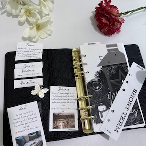 May include: A black leather planner with gold-colored rings, open to reveal inserts with text and images. Includes cards with words like "Peace," "Growth," and "Balance." A decorative butterfly pin and a "Short Term" tab are also visible.