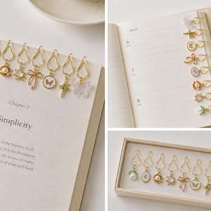 May include: Gold-toned paper clips with decorative charms, including flowers, pearls, and butterflies, adorn an open book. The text "Simplicity" is visible. A set of clips is also displayed in a box, and some are clipped to a planner.