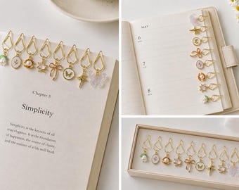 Puede incluir: Clips dorados con colgantes decorativos, incluyendo flores, perlas y mariposas, adornan un libro abierto. El texto "Simplicity" es visible. Un conjunto de clips también se muestra en una caja y en una agenda.