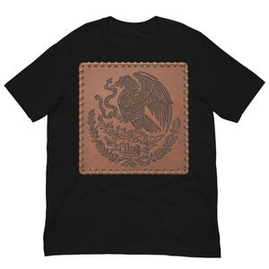 Può includere: Una t-shirt nera a maniche corte con una grafica rettangolare centrale che imita una toppa in pelle marrone. La toppa presenta un disegno in rilievo di un'aquila appollaiata su un cactus che tiene un serpente, circondata da rami di alloro.