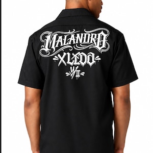 May include: A black short-sleeved button-down shirt featuring white graphic text on the back. The design includes the word MALANDRO in a stylized font, followed by XLIDO and the Roman numeral VII, all rendered in a bold, white, graffiti-inspired script.
