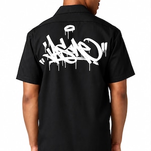 May include: A black short-sleeved button-down shirt featuring a large white graffiti-style graphic on the back. The design consists of stylized, dripping white lettering and a circular symbol above the text, centered on the upper back area.