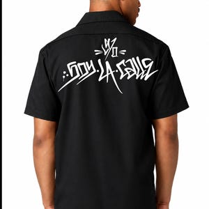 May include: A black short-sleeved button-down shirt featuring a white graffiti-style graphic on the back. The design includes stylized lettering that reads "SOY DE LA CALLE" with an abstract symbol above the text, printed in a bold white ink.