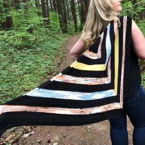 May include: A woman wearing a black tank top and blue jeans stands in a forest. She is wearing a black, brown, yellow, and blue striped shawl.