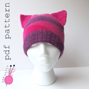 May include: A pink and purple striped knit hat with cat ears. The hat is on a white mannequin head.