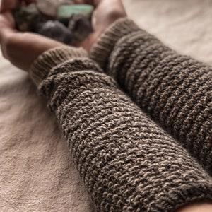 Puede incluir: Primer plano de un par de calentadores de brazo de punto marrón y beige. Los calentadores de brazo están hechos de un hilo suave y texturizado y tienen un patrón de canalé.