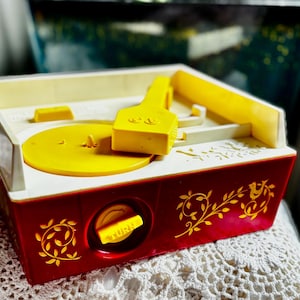 May include: A vintage toy record player with a red base and yellow accents. The turntable, arm, and controls are yellow, while the top and sides are white. The red base features gold floral and bird designs.