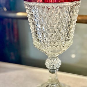 May include: A clear glass goblet featuring a diamond-cut pattern on the bowl and a deep red rim. The stem includes a textured, faceted knob detail, and the base is circular and smooth. The glass reflects light across its geometric surface.