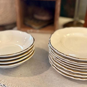 May include: A set of white ceramic bowls and plates with gold rims. The bowls have a scalloped edge and are stacked in a neat pile. The plates are also stacked, showcasing a similar design. The tableware is likely for serving food.