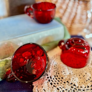 May include: Three ruby red glass teacups with handles. The cups are on a white crocheted doily and a blue book. The cups are a deep, transparent red, reflecting light. The teacups are a vintage style.
