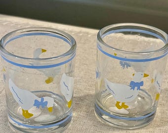 May include: Two clear glass shot glasses featuring a repeating pattern of white geese with yellow beaks and feet, wearing blue bows around their necks. Each glass is decorated with thin horizontal light blue stripes at the top and bottom rims.