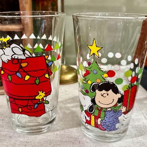 May include: Two clear glass tumblers with Christmas-themed cartoon graphics. One features Snoopy on his doghouse with Christmas lights, the other Lucy with a Christmas tree and presents. Both have white, green, and red accents.