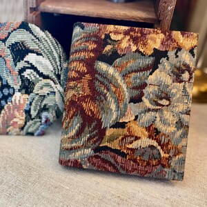 May include: Two decorative items with floral tapestry patterns. One is a round, padded object with a blue, green, and pink floral design. The other is a rectangular, padded object with a burgundy, gold, and blue floral pattern.