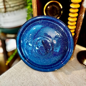 May include: A blue enamelware lid with white speckles. The lid has a rounded shape with a central handle. The lid is sitting on a neutral-colored linen tablecloth. The lid is part of a camping or outdoor cooking set.