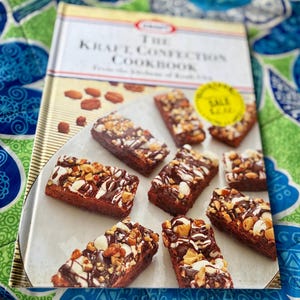 May include: A cookbook titled "The Kraft Confection Cookbook" with a photo of brownies topped with nuts and chocolate drizzle. The brownies are arranged on a white plate. A yellow "SALE" sticker is visible.