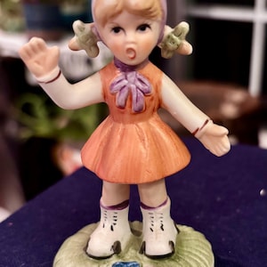 May include: A ceramic figurine of a child with blonde hair wearing a purple bonnet, an orange dress with a purple bow, and white boots. The child stands on a green base with small blue and pink flowers, with one arm raised and mouth open.
