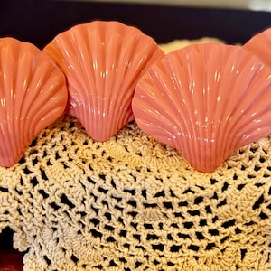 May include: Four coral-colored, ceramic seashell-shaped dishes are arranged on a cream-colored, crocheted doily. The shells have a ribbed texture and are evenly spaced, creating a decorative display.