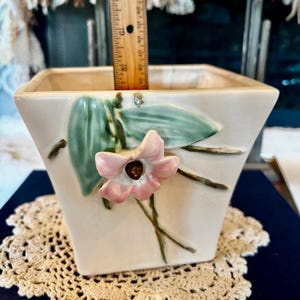 May include: Cream-colored ceramic square planter with a raised floral design. The planter features a pink flower with a brown center and green leaves. A ruler shows the planter is approximately 5 inches tall.