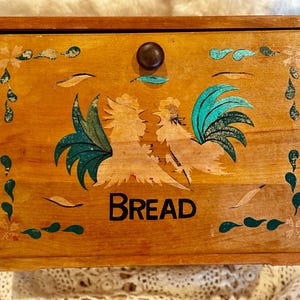 May include: Wooden bread box with a rooster design. The box is rectangular with a dark brown knob and the word "BREAD" in black letters. The rooster has teal and white accents. The box has decorative teal and white accents.