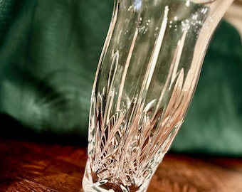 May include: A clear, cut-glass vase with a flared top and a detailed, etched pattern. The vase has a slightly curved shape and sits on a wooden surface. The background features a dark green fabric.