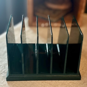 May include: A black acrylic desk organizer featuring five vertical slots for holding documents or mail. The structure has a rectangular base and upright dividers with angled top corners, creating a clean, multi-compartment storage unit.