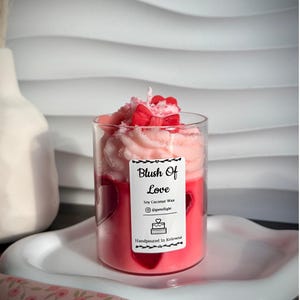 Può includere: Una candela di vetro con cera rosa e rossa, sormontata da un vortice di cera rosa e un fiocco rosso. La candela ha un'etichetta che recita "Blush Of Love" ed è versata a mano a Kelowna. Poggia su un piatto bianco a forma di cuore.