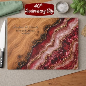 May include: A rectangular wooden cutting board featuring a natural wood grain on the left and a decorative red, pink, and gold resin geode-style inlay on the right. The text "Andrew & Nicole Forever & Always 40 YEARS" is engraved on the wood surface.