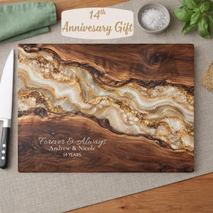 May include: A rectangular wooden cutting board featuring a central resin inlay with white and gold marbled patterns. The board is engraved with the text "Forever & Always", "Andrew & Nicole", and "14 YEARS". A white knife is positioned to the left.