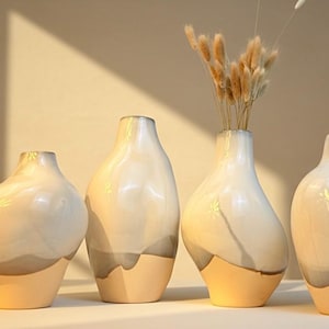 May include: Four ceramic vases with a glossy finish. The vases are white with a beige base and a gray accent. One vase holds dried ornamental grass. The vases have a unique, organic shape, perfect for home decor.