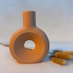 May include: A matte peach-colored ceramic vase with a circular, donut-shaped body featuring a central hollow opening and concentric ribbed detailing. The vase has a cylindrical neck and a flat base, accompanied by dried beige grass stems.