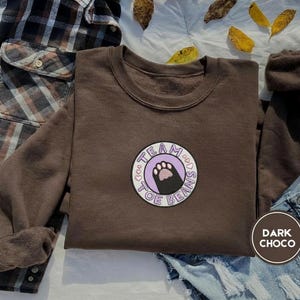 May include: Dark chocolate brown crew neck sweatshirt with a circular patch featuring a paw print and the words "Team Toe Beans". The sweatshirt is displayed with a plaid shirt and denim shorts.