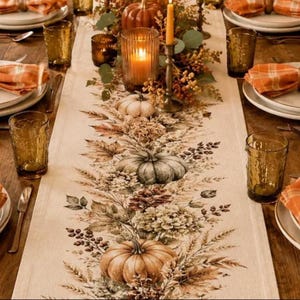May include: A cream-colored table runner featuring a printed design of pumpkins, autumn leaves, pinecones, and wheat stalks. The runner is set with amber-colored glassware, orange plaid napkins, and a central candle arrangement.
